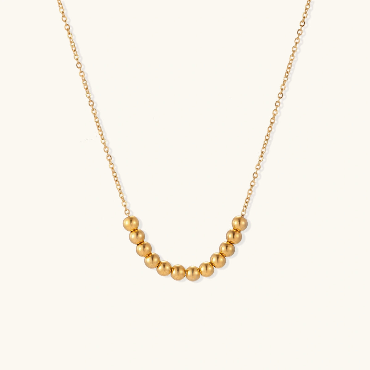 Delicate gold-plated necklace with graduated spherical beads forming gentle smile curve on beige background